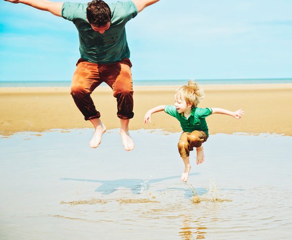 A white man and white boy, both wearing green t-shirts and brown trousers, jumping in mid air above a pool of water on a sandy beach. 